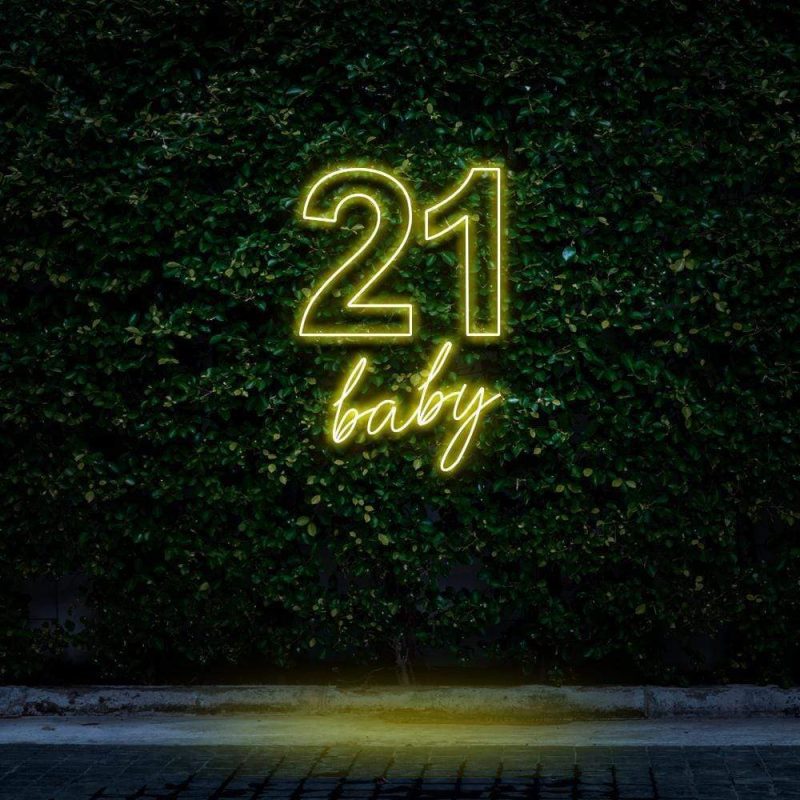 Bright yellow neon sign reading "21 Baby," glowing against a green leafy wall, ideal for 21st birthday celebrations and festive party decorations.